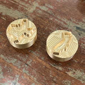May include: Two gold-colored metal stamps with a raised design of a stylized arrow shape. The stamps are circular and have a smooth, polished surface.