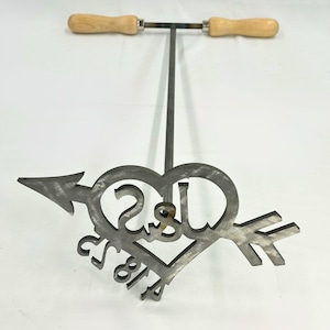 May include: A metal heart-shaped branding iron with an arrow through it. The heart has the letters "J & S" and the numbers "5.8.14" inside. The iron is attached to a long metal rod with wooden handles.