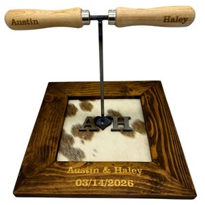 May include: A personalized wooden branding iron with the names "Austin" and "Haley" on the handles. The iron is mounted on a wooden frame with a cowhide background, featuring the initials "A" and "H" joined by a heart, and the date "03/14/2026".