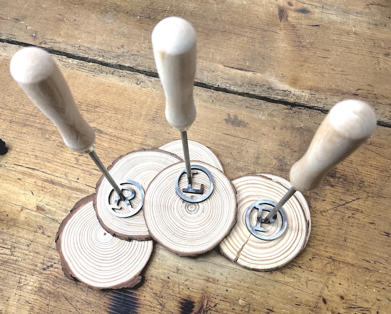 Op de afbeelding: Drie brandingijzers met houten handvatten en metalen punten in de vorm van de letters R, L en E. De ijzers worden gepresenteerd op drie houten rondjes.