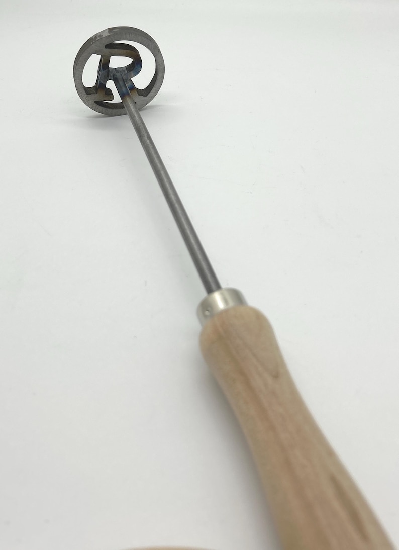 Op de afbeelding: Een metalen brandmerk met een houten handvat. Het brandmerk heeft een ronde kop met de letter "R" uitgesneden.