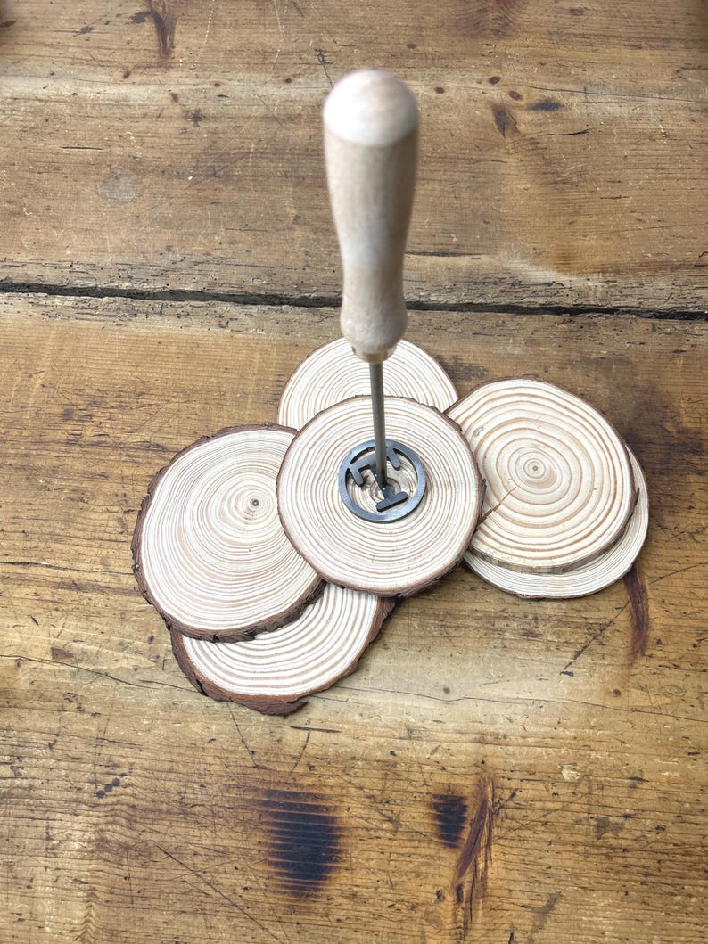 Op de afbeelding: Een set van vier houten onderzetters met een metalen brandmerk in het midden. De onderzetters zijn gemaakt van natuurlijk hout met een lichtbruine kleur en hebben een ronde vorm. Het brandmerk is een eenvoudig ontwerp met een enkele letter "T" in het midden.