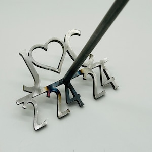 May include: A metal branding iron with the letters "J &" and a heart, followed by the numbers "2424". The branding iron is attached to a metal rod.