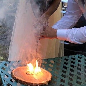 Op de afbeelding: Een stel voert een vuurceremonie uit met behulp van een handboor om houtsnippers op een houten schijf te ontsteken. Rook en vlammen stijgen op uit het hout terwijl ze samenwerken.