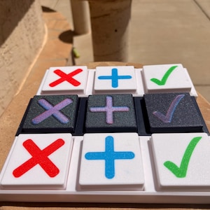 May include: A square game board with a three-by-three grid containing nine tiles. The tiles feature red X symbols, blue plus signs, and green check marks. Some tiles are white with coloured symbols, while others are dark grey with iridescent symbols.