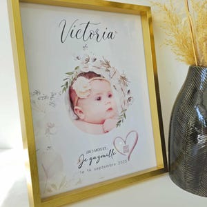 Peut inclure: Un cadre doré encadre une photo de bébé nommée Victoria, ornée d'une couronne florale. Le texte "J'ai 3 mois et... Je gazouille le 14 septembre 2025" est inclus. Un vase décoratif est visible en arrière-plan.