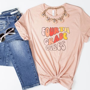 May include: A pink t-shirt with the text "FOURTH GRADE VIBES" in a rainbow color scheme. The text is in a retro font style.