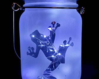 May include: A frosted glass jar with a metal lid and a wire handle. The jar has a silhouette of a frog with a string of lights inside.