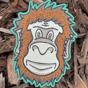 Puede incluir: Un parche bordado con la cara de un orangután, con una expresión amigable, en marrón y verde.