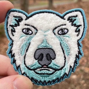 以下が含まれることがあります： 青い縁取りと青いグレーのディテールで刺繍された白い犬の顔のアップリケ。犬は白い毛並みのテクスチャで、青いアウトラインがあります。