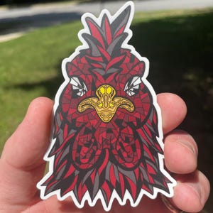 May include: A sticker featuring a stylized red, black, and gray bird head with a gold-colored emblem on its chest. The bird's head is intricately designed with geometric patterns and a white border.