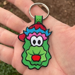 May include: Embroidered keychain featuring a whimsical green character with a red hat, blue and pink accents, and large black and white eyes. The keychain includes a metal ring and a green fabric tab with the letters "RY".