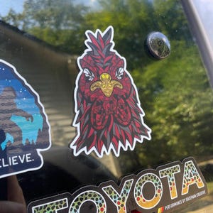 May include: A sticker featuring a stylized rooster head with a red and gray mosaic design. The rooster has a gold beak and is set against a white border. The sticker is affixed to a dark surface.