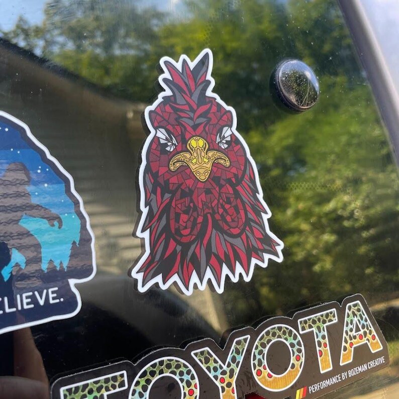 May include: A sticker featuring a stylized rooster head with a red and gray mosaic design. The rooster has a gold beak and is set against a white border. The sticker is affixed to a dark surface.