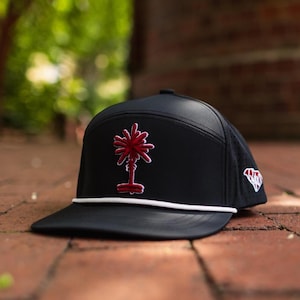 May include: A black baseball cap featuring a red embroidered palmetto tree emblem on the front panel. The cap includes a white rope detail across the base of the crown and a small white and red embroidered state outline on the side panel.