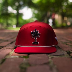 May include: A deep red baseball cap featuring a structured crown and a flat brim. A black embroidered palm tree graphic with a white outline is centered on the front panel. A thin white cord runs horizontally across the base of the crown.