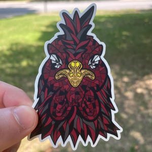 May include: A sticker featuring a stylized rooster head with a mosaic-like design. The rooster is primarily red and black, with a gold-colored beak and accents. The sticker has a white border and is held against a blurred green background.