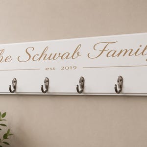 May include: A rectangular white wooden wall-mounted coat rack featuring six silver metal hooks. The text "The Schwab Family" is printed in a brown script font above the text "est 2019" which is centered between two horizontal brown lines.