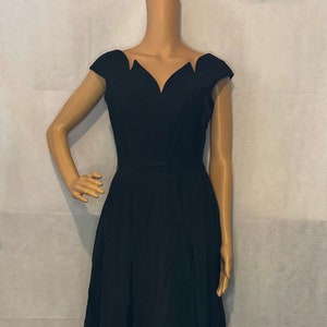 May include: A black dress with a unique neckline and cap sleeves. The dress has a fitted bodice and a flared skirt that falls to the knees. The dress is displayed on a mannequin.