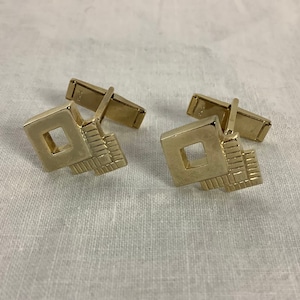 May include: A pair of gold-toned cufflinks with a geometric design. Each cufflink features a square with a smaller square cut out, and a rectangular base with a series of parallel lines. The cufflinks are on a white surface.