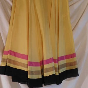 May include: A long, flowing yellow skirt with a pleated waist. The skirt features horizontal bands of pink, brown, and black near the hem. The fabric appears lightweight and suitable for warm weather.