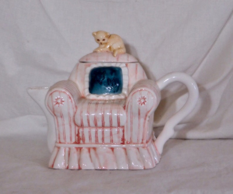 May include: A ceramic teapot shaped like a pink and white striped armchair, with a small beige cat figurine on the lid. The chair has a blue square with a floral design. The teapot has a white handle and spout.