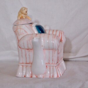 May include: A white ceramic teapot shaped like a striped armchair with red accents. A small, tan dog figurine sits on the lid. A white creamer is in front of the chair, and a blue pillow is on the chair.