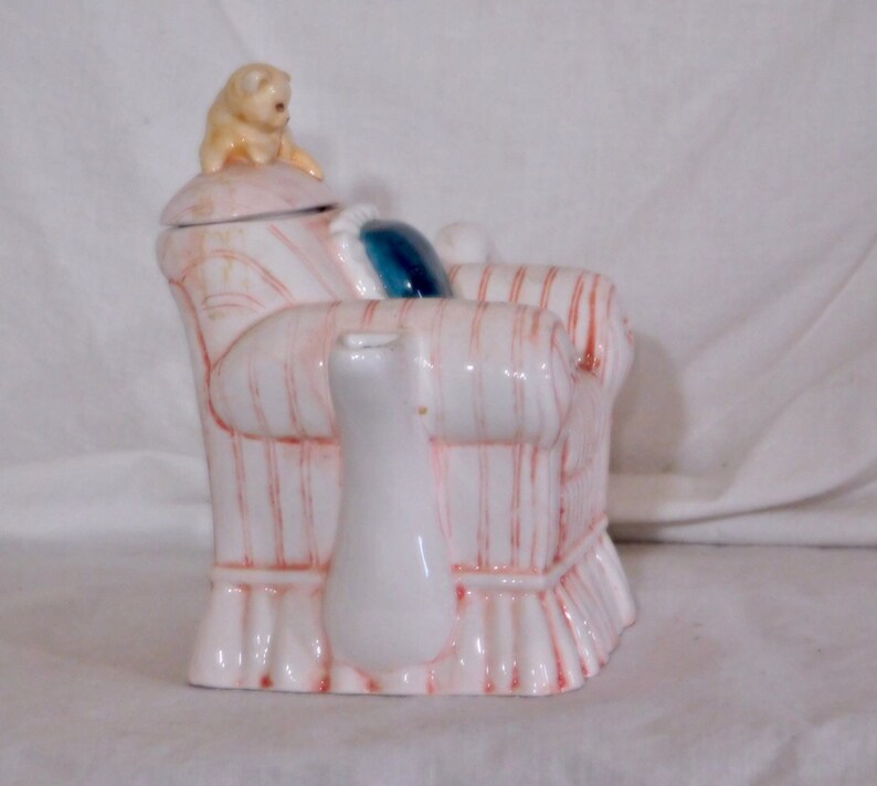 May include: A white ceramic teapot shaped like a striped armchair with red accents. A small, tan dog figurine sits on the lid. A white creamer is in front of the chair, and a blue pillow is on the chair.