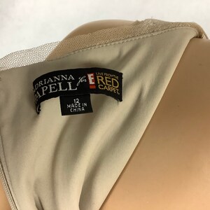 May include: Close-up of a beige dress with a delicate lace neckline. The label reads "Adrianna Papell for the Red Carpet" and indicates a size 12, made in China. The dress is displayed on a mannequin.