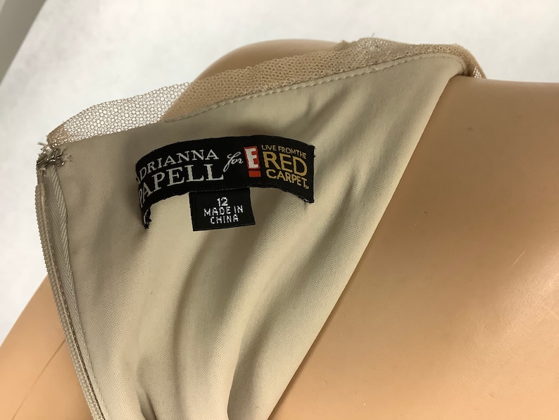 May include: Close-up of a beige dress with a delicate lace neckline. The label reads "Adrianna Papell for the Red Carpet" and indicates a size 12, made in China. The dress is displayed on a mannequin.