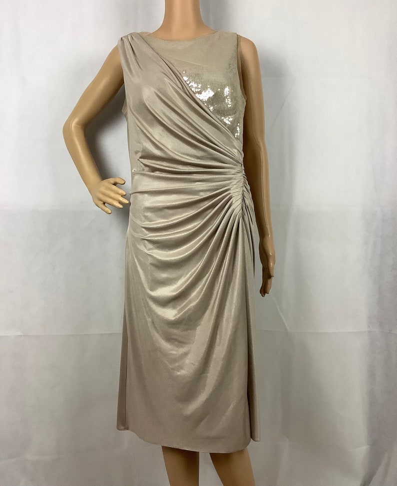 May include: A sleeveless, knee-length dress in a champagne color. The dress features a draped design with ruching on the side and a sequined panel on the bodice. The dress is displayed on a mannequin.