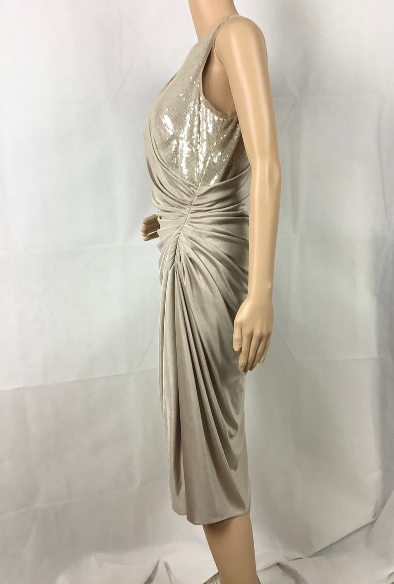 May include: A beige, sleeveless dress with a knee-length hemline. The bodice features a sequined panel, and the dress has a draped, ruched design. The dress is displayed on a mannequin against a white background.