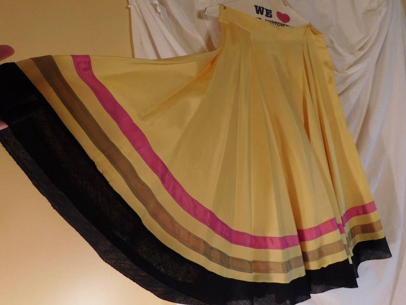 May include: A flowing yellow skirt with a wide, striped hem. The stripes include black, brown, pink, and tan. The skirt is made of a lightweight fabric and is hanging on a hanger.