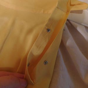 May include: Close-up of a yellow garment with silver snap closures. The fabric appears to be lightweight and slightly translucent. The garment is partially open, revealing the interior construction and the snap details.