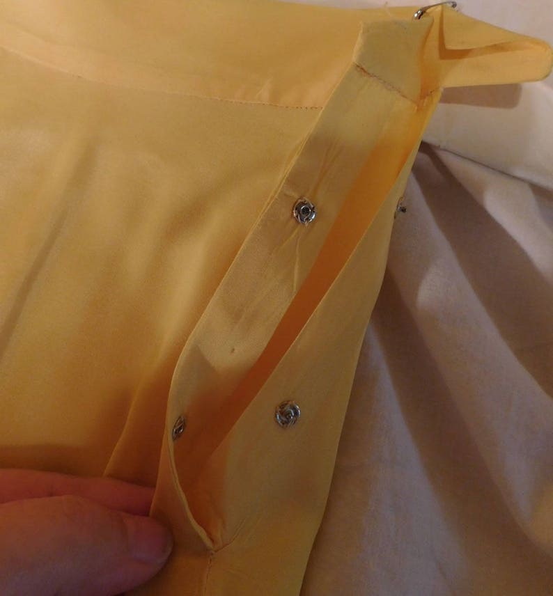 May include: Close-up of a yellow garment with silver snap closures. The fabric appears to be lightweight and slightly translucent. The garment is partially open, revealing the interior construction and the snap details.