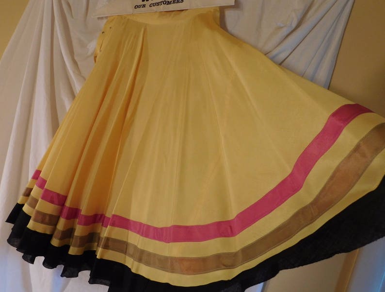 May include: A flowing, yellow skirt with a wide, flared design. The skirt features horizontal stripes of pink, brown, and black near the hem. The fabric appears lightweight and has a slight sheen.