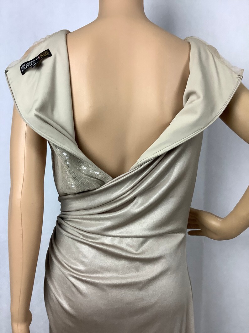 May include: A beige, draped evening gown with a cowl neckline and a low back. The dress features a sequined panel and a zipper closure. The label reads "Adrianna Papell". The dress is displayed on a mannequin.