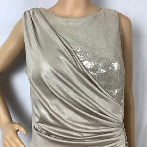 May include: A sleeveless, champagne-colored dress with a draped bodice and a sequined panel. The dress features a ruched detail on the side, creating a flattering silhouette. The fabric has a subtle sheen, adding to its elegant appearance.