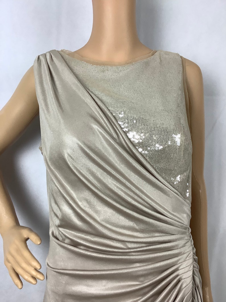 May include: A sleeveless, champagne-colored dress with a draped bodice and a sequined panel. The dress features a ruched detail on the side, creating a flattering silhouette. The fabric has a subtle sheen, adding to its elegant appearance.