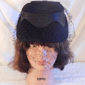 May include: A black hat with a rounded crown and a decorative band. The hat features a delicate black netting veil that drapes over the face. The hat is displayed on a mannequin head.