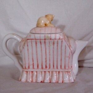 May include: A white ceramic teapot with a lid and handle. The teapot is decorated with vertical red stripes and a small, light brown bear figurine on top. The base has a ruffled design.