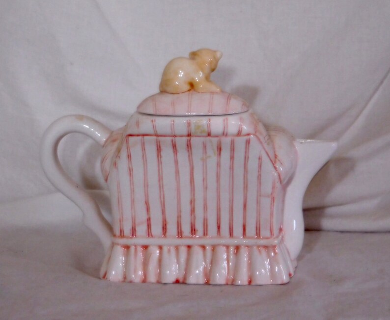 May include: A white ceramic teapot with a lid and handle. The teapot is decorated with vertical red stripes and a small, light brown bear figurine on top. The base has a ruffled design.