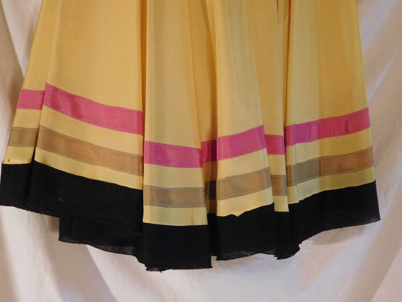 May include: A flowing, yellow skirt with horizontal stripes of pink, tan, and black. The skirt appears to be made of a lightweight fabric, with a slight sheen. The hemline is finished with a wide black band.