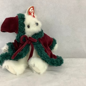 May include: A white plush polar bear wearing a burgundy velvet coat with green trim and a matching hood. The coat is tied with a burgundy bow. A red tag is attached to the bear's ear. This stuffed animal is a festive holiday decoration.