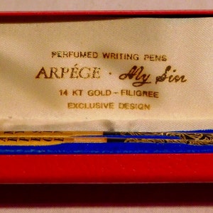 May include: A vintage pen set in a red and blue velvet case. The case is open, revealing a gold-colored pen with black filigree and the text "ARPEGE - My Sin". The case also has the text "PERFUMED WRITING PENS" and "14 KT GOLD - FILIGREE".