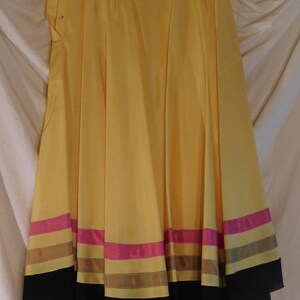 May include: A flowing, yellow skirt with a wide hemline. The skirt features horizontal bands of pink, brown, and black near the bottom. The skirt is made of a lightweight fabric and has a button closure at the waist.
