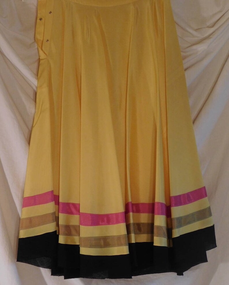 May include: A flowing, yellow skirt with a wide hemline. The skirt features horizontal bands of pink, brown, and black near the bottom. The skirt is made of a lightweight fabric and has a button closure at the waist.
