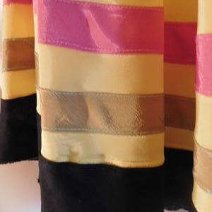 May include: Close-up of several fabric columns with horizontal stripes. The columns are primarily yellow with pink and brown stripes. The bottom of each column is black with a lace trim.