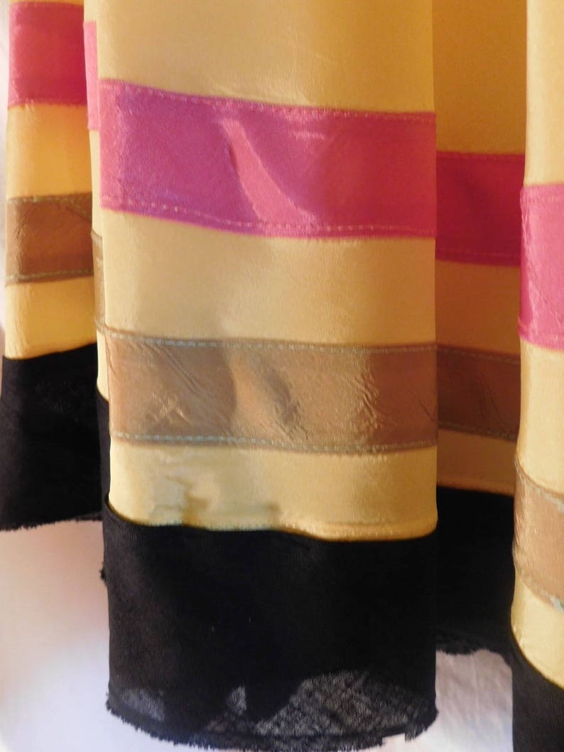 May include: Close-up of several fabric columns with horizontal stripes. The columns are primarily yellow with pink and brown stripes. The bottom of each column is black with a lace trim.