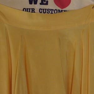 May include: A flowing, yellow skirt with a wide waistband. The fabric drapes in soft folds, creating a full silhouette. The skirt is suspended from a metal clip. The background includes the text "WE OUR CUSTOM".